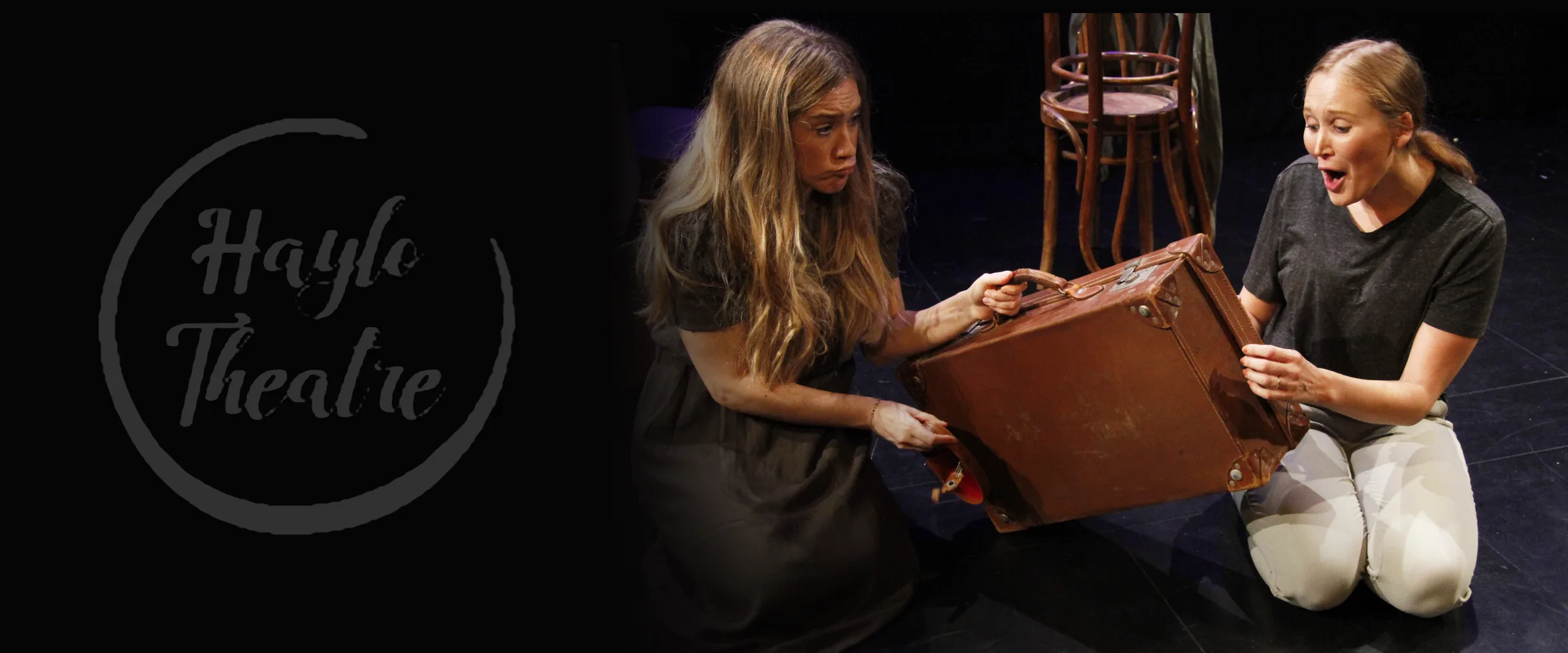 Two women kneel beside a brown suitcase on a dark stage, with stacked chairs behind them. A circular “Haylo Theatre” logo sits to the left.
