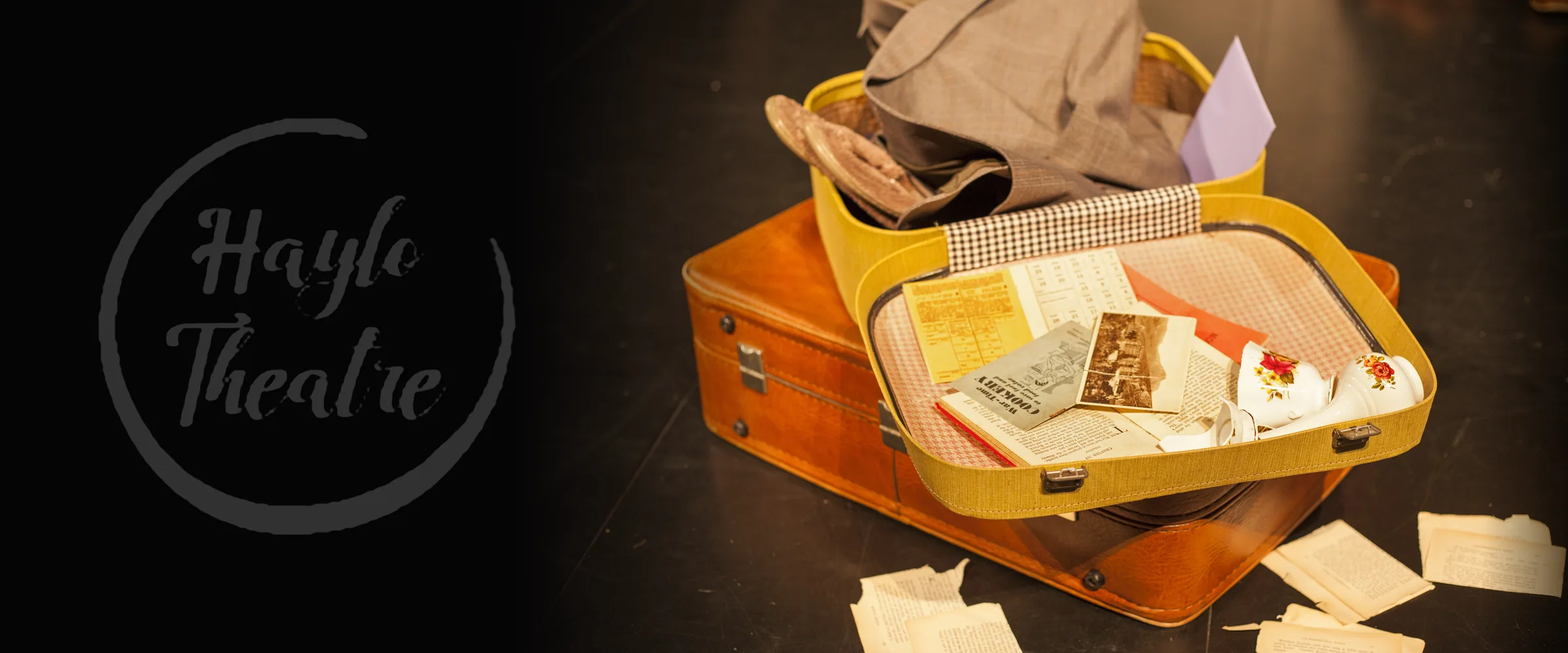 An open yellow suitcase holds floral cups, photographs and books atop a larger brown case. Text: “Haylo Theatre”; “War-Time COOKERY to save fuel and food value”. Smaller printed text is illegible.