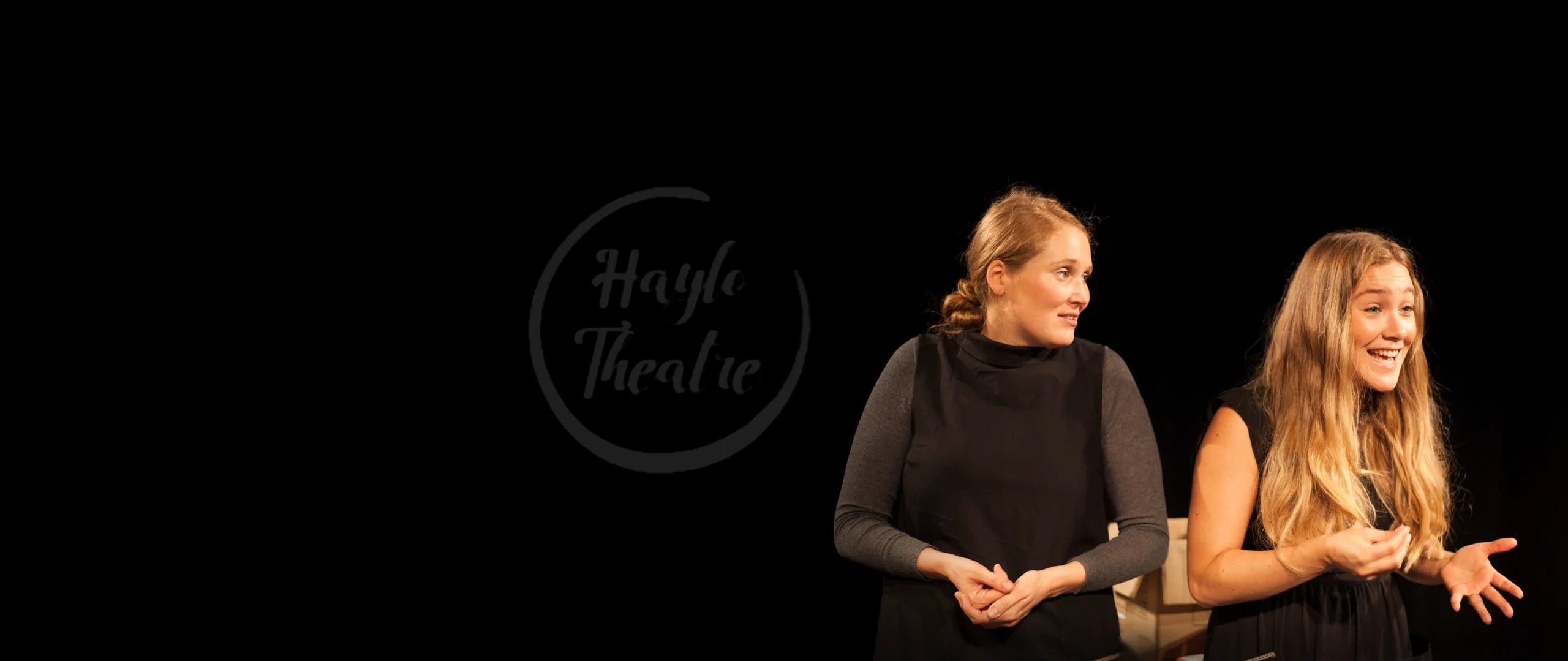 Two women in black gesture during a performance against dark curtains; a circular “Haylo Theatre” logo sits on the black left side.