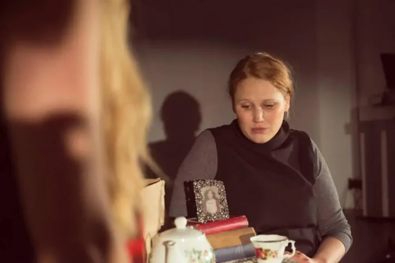 A seated woman looks down towards a tea set beside stacked books and a framed portrait; another woman is blurred in the foreground.