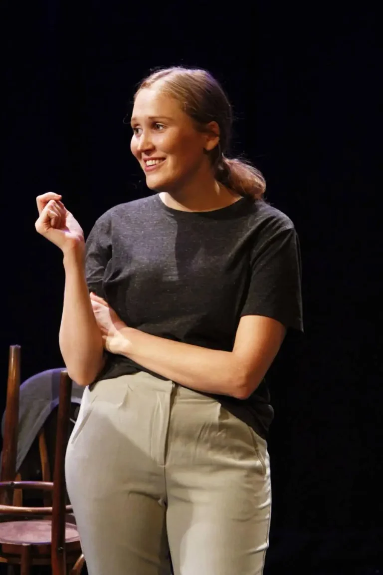 A woman in a black T-shirt and grey trousers gestures with one hand near her shoulder and the other across her waist on a dark stage.