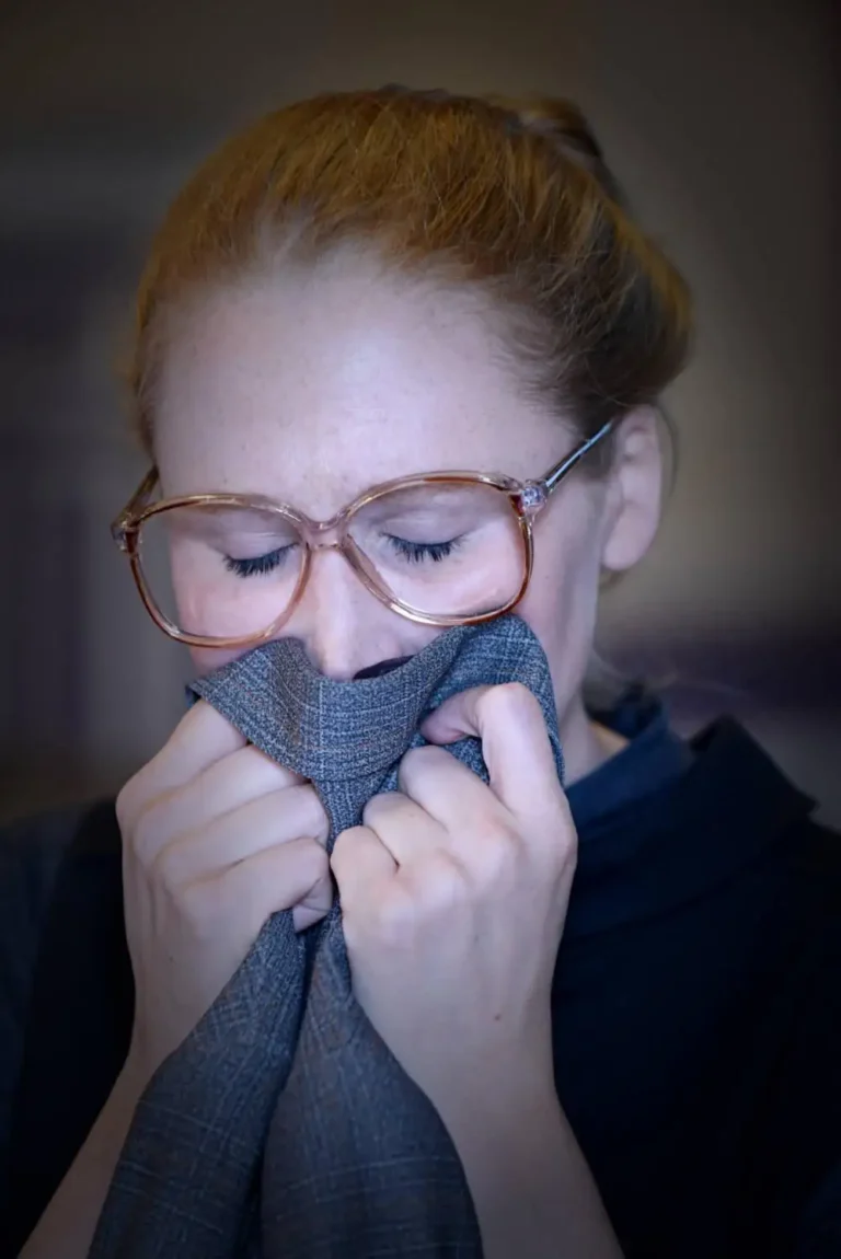 A woman wearing glasses holds grey fabric over her mouth and nose with both hands, her eyes closed during a performance.
