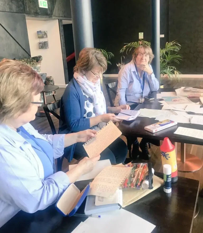 Three women sit around a table examining papers and notebooks among drawing materials and glue. A container partly reads “GLU… STI…”. Handwriting and remaining small print are illegible.