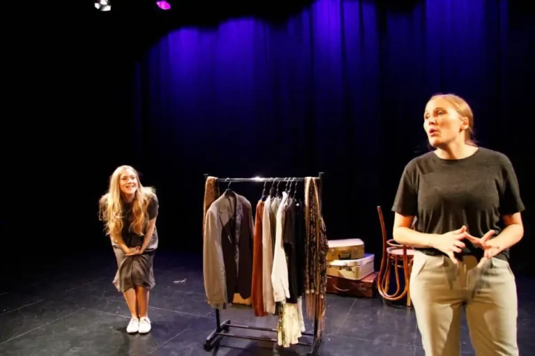 Two women in black perform beside a clothes rail; one gestures in the foreground while the other smiles and leans forward against blue-lit curtains.