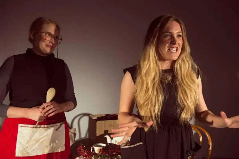 A woman gestures with open palms while another, wearing glasses and a red apron, holds a wooden spoon beside a table of stage props.