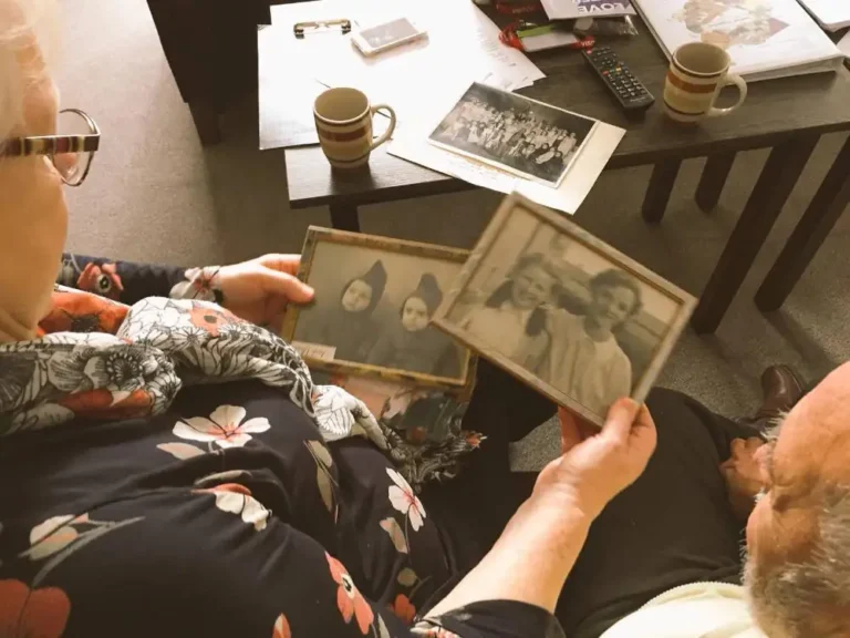 An older woman holds two framed photographs beside an older man at a table with mugs and papers. Visible text: “LOVE”. Smaller labels and print are illegible.
