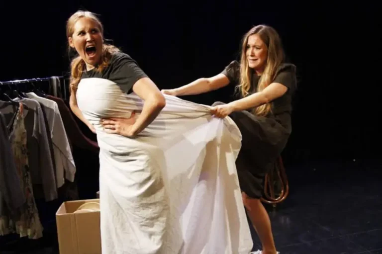 A woman stands wrapped in a white sheet with one hand on her hip while another woman holds the fabric behind her on a stage.